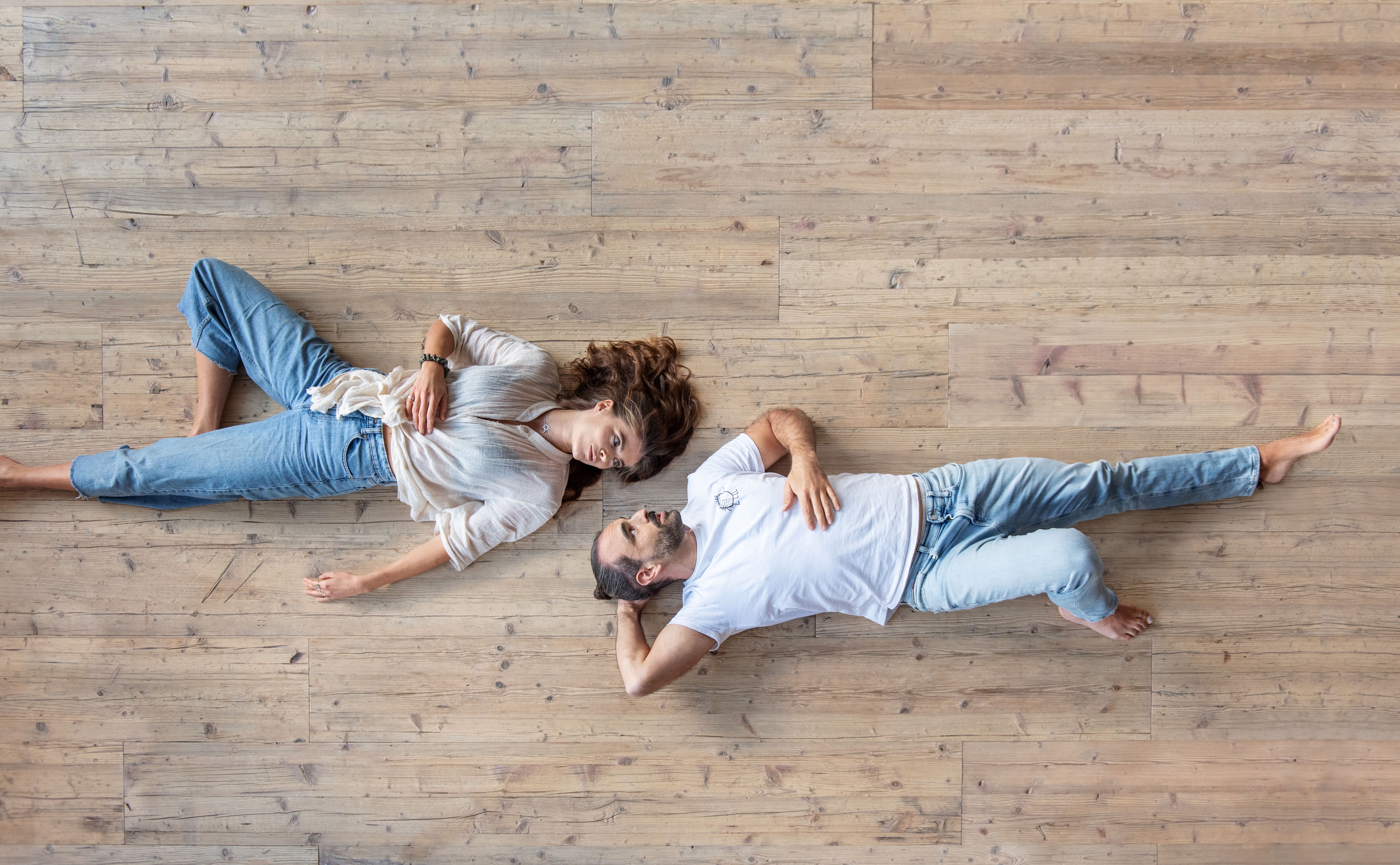 Eine Frau und ein Mann liegen entspannt auf dem Rücken auf einem Boden aus Altholzdielen und sehen sich an.