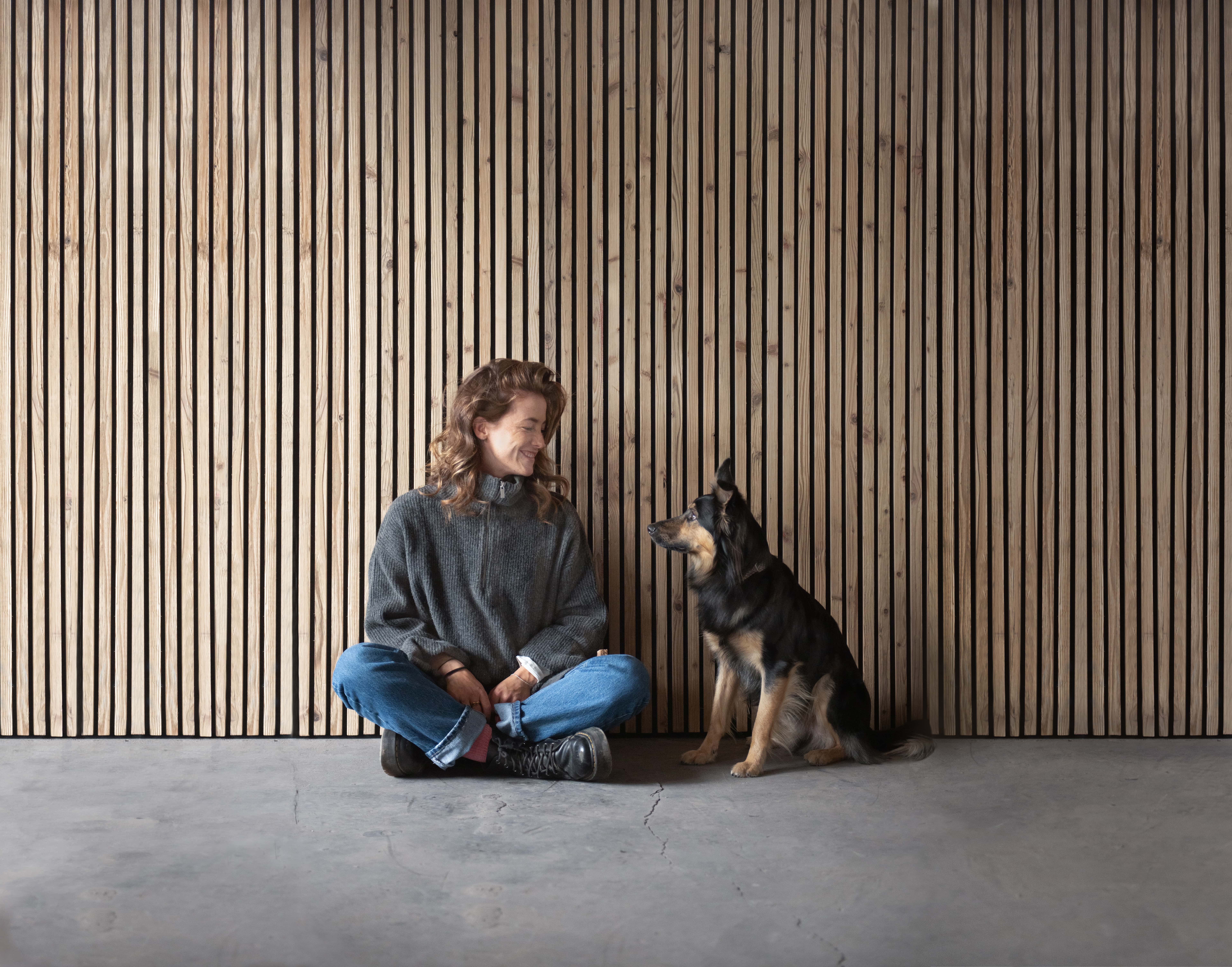 Eine lächelnde, junge Frau und ein Hund sitzen vor einer mit Akustikpaneelen aus Altholz verkleideten Wand und sehen sich an.